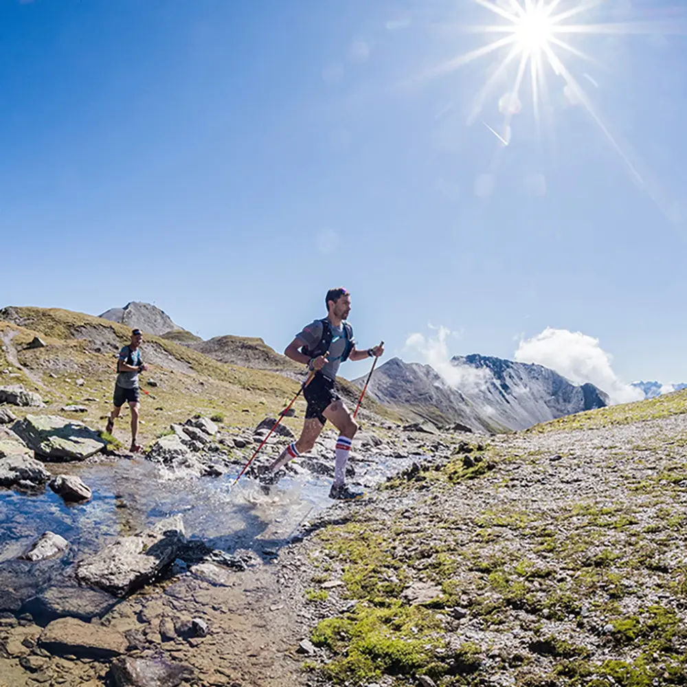 Zwei Personen beim Trailrunning mit Stöcken über einen felsigen Bach in den sonnigen Samnauner Bergen.