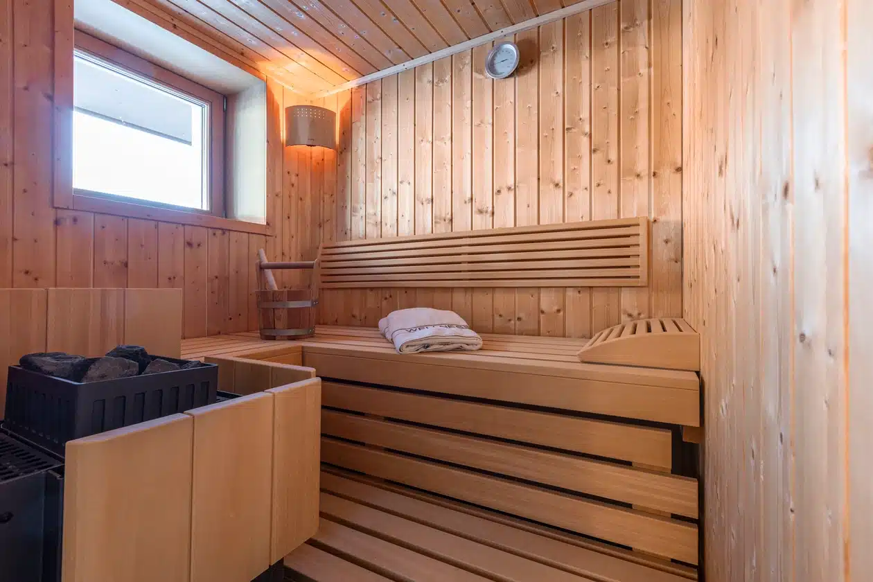 Hölzerne Saunakabine mit Sitzbank, Handtüchern, einem Eimer und einem Fenster, das Tageslicht hereinlässt.