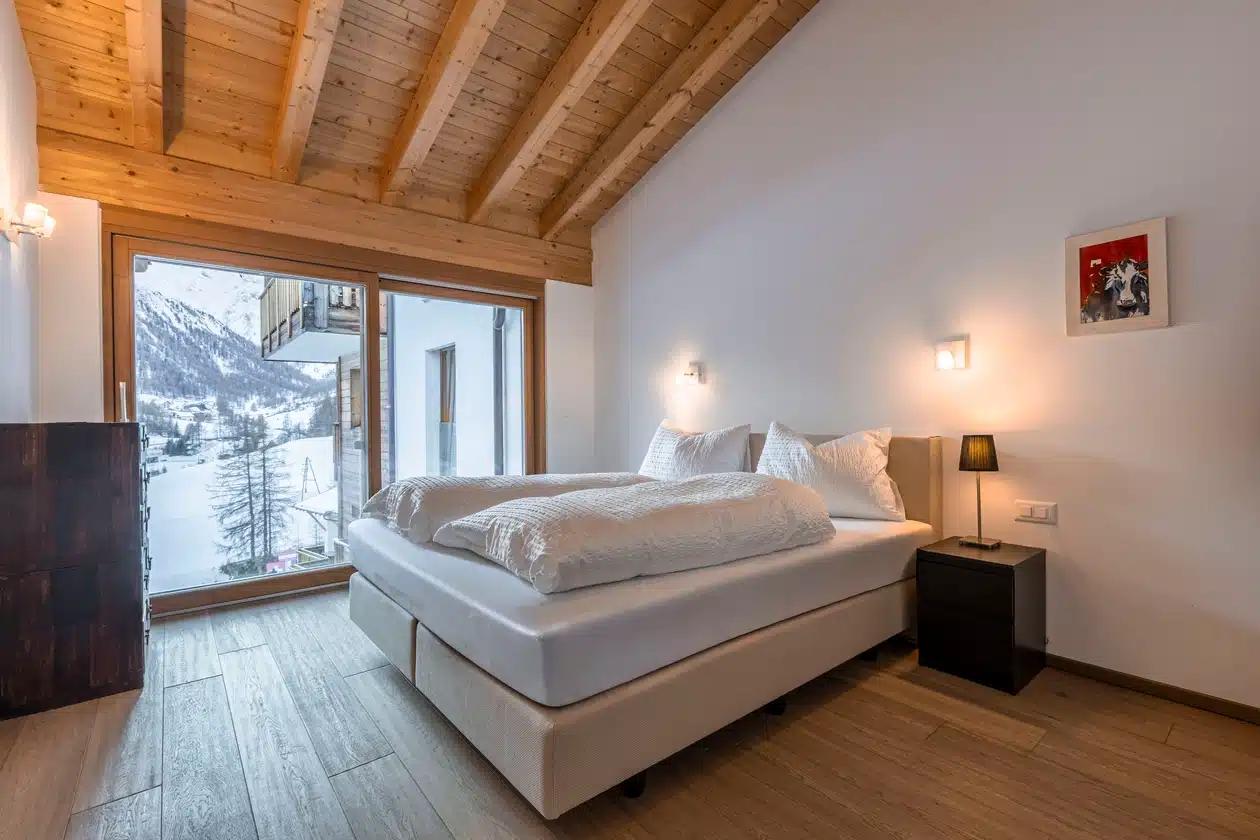 Modernes Schlafzimmer mit großem Bett, Holzdecke und einem Fenster mit Blick auf die verschneiten Berge.