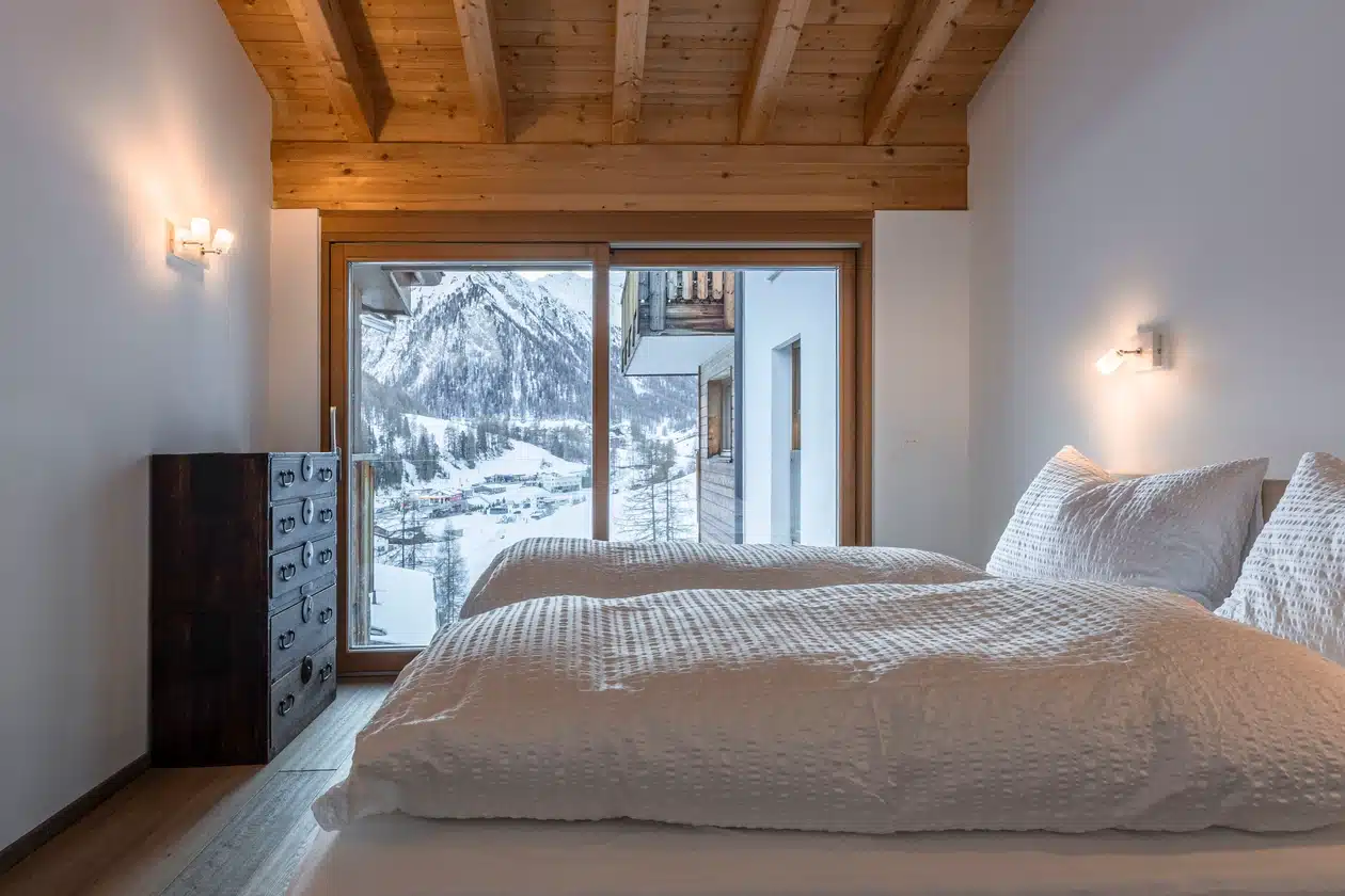 Gemütliches Schlafzimmer mit zwei Einzelbetten, Holzdecke, großem Fenster und Blick auf die verschneiten Berge draußen.