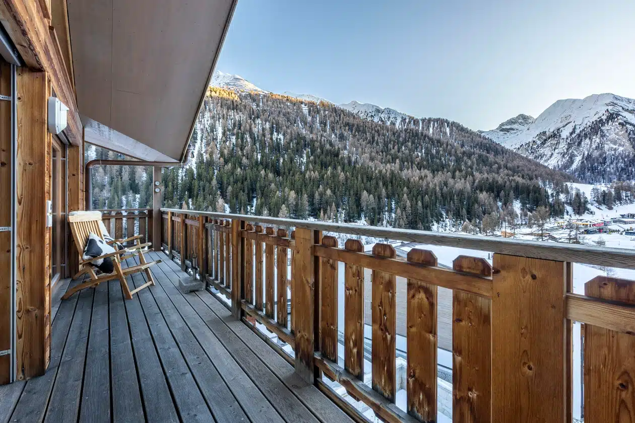 Holzbalkon mit Stühlen und Blick auf die schneebedeckten Berge und Kiefern.
