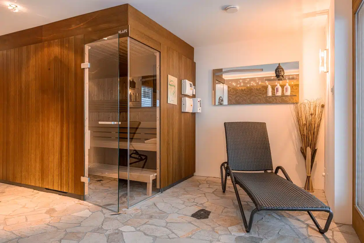 Eine moderne Innensauna mit einer Glastür und einem schwarzen Sessel auf einem Steinboden.