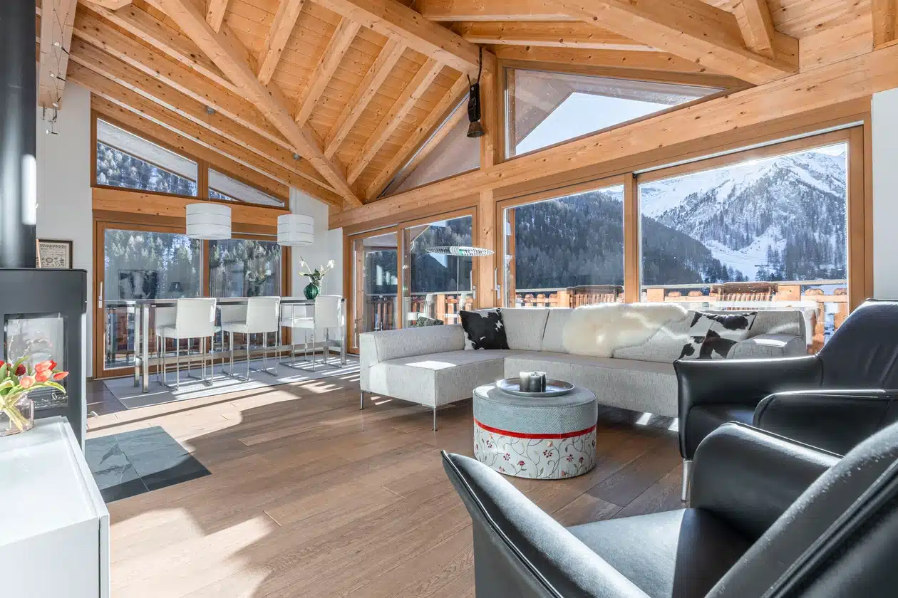 Modernes Wohnzimmer mit Holzbalken, großen Fenstern und Blick auf die Berge in einer verschneiten Umgebung.