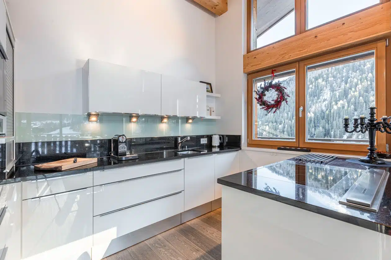 Moderne weiße Küche mit schwarzen Arbeitsplatten, großem Fenster und Blick auf die Berge.